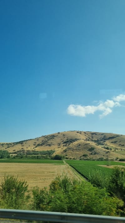 The image shows a landscape scene with a clear blue sky dominating the upper portion. A single, wispy white cloud is visible in the sky. Below the sky, a large, dry, golden-brown hill covered in sparse vegetation stretches across the midground. In the foreground, there are agricultural fields. To the left, a field appears to have been harvested, with dry, straw-like residue. To the right of this, a vibrant green field, possibly a vineyard or crop of young plants, is neatly organized in rows. A few scattered trees and bushes dot the landscape between the fields and the hill. In the very front, a blurred metal guardrail runs horizontally, suggesting the photo was taken from a moving vehicle. The overall impression is a rural, agricultural landscape under a bright sky. The prompt requests a description including the location in the city of Municipal Unit of Kilada, Greece. However, there are no specific visual cues in the image that would allow for a precise identification of this location. The scene is generic of a hilly, agricultural region.