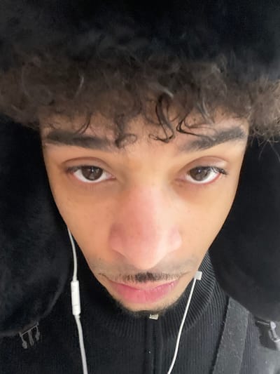 A close-up, high-angle photograph depicts a single male individual with medium brown skin and dark, curly hair visible at the forehead. The individual wears a dark, likely black, furry or plush hat with ear flaps pulled down, framing the face. His dark brown eyes are open and directed at the camera. A thin mustache and light stubble are present on the chin. The subject is dressed in a dark, high-collared, ribbed zippered garment. White wired earbuds are inserted into both ears, with a small control unit visible on the wire descending from the individual's left ear. The background is uniformly dark, offering no specific environmental details within the frame. The scene is located in the city of Plaisir, France.