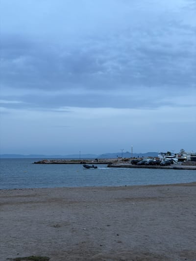 The image shows a cloudy sky above a harbor in Glifada, Greece. The harbor has a breakwater wall made of rocks with a small boat in the water near it. Parked vehicles and buildings are visible along the waterfront. A sandy beach is present to the right of the harbor.