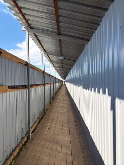 The image depicts a long, covered walkway with a wooden floor and corrugated metal walls. The walkway is enclosed by fences on both sides, with a metal roof supported by a metal frame. The sky is visible through gaps in the roof and to the left of the walkway. The sun casts shadows on the right-hand wall, creating a pattern of vertical lines. A single person is visible walking away from the camera at the far end of the walkway. The environment appears to be outdoors, possibly in an industrial or construction area, given the materials used. The image was taken in Saint Petersburg, Russia.