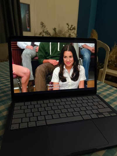 The image captures a tablet, likely an iPad, displaying a video, positioned on a blue and white checkered tablecloth. The tablet is attached to a dark-colored keyboard case, which is open to the right.
On the tablet screen, a woman with dark hair, wearing a white collared shirt, is prominently featured, smiling towards the viewer. She appears to be an actress in a scene from a TV show or movie. Parts of other people are visible around her on the screen, including one individual in a green top and another in grey. Subtitles are displayed on the lower right of the screen, reading: "* Who'd have believed you'd come along *".
The keyboard case shows a light grey QWERTY key layout, including Spanish characters (e.g., 'Ñ'). Visible keys include 'control', 'option', 'command', 'shift', 'tab', 'esc', 'caps lock', 'fn', and various letter, number, and symbol keys. A dark trackpad is integrated into the right side of the keyboard.
The background is dimly lit, suggesting an indoor setting during the evening or night. Blurred elements include furniture, such as a chair, and indistinct decorative items that resemble plants. The scene takes place in Urbanización Encinares De Alcolea, Spain.