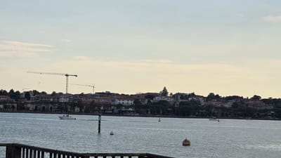 The image depicts a serene waterscape with a town nestled along the coastline in the background. Two large construction cranes dominate the skyline, suggesting ongoing development. The water is calm, with a few boats visible, including a white speedboat carrying people. Several buoys dot the water's surface, marking navigational channels or areas. The town itself is characterized by a collection of buildings with terracotta roofs, interspersed with trees. The sky is overcast with diffused light, indicating either early morning or late afternoon. The overall mood is tranquil, with a hint of industrial activity due to the presence of the cranes. The location is identified as Peschiera del Garda, Italy, a setting known for its picturesque views and historical significance.