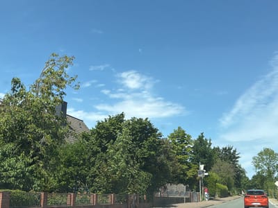 The image shows a street scene in Dobersdorf, Germany, on a sunny day with a clear blue sky and scattered white clouds. A red car is driving away from the viewer on a paved road that curves to the right. The car has German license plates. To the left of the road, there is a brick fence with decorative metal inserts, behind which is a lush green garden with various trees and bushes. A building with a grey tiled roof and a chimney is partially visible behind the trees. Further down the road, there are more trees and some street furniture, including a signpost and a bench. The overall impression is of a quiet, residential area on a summer day.