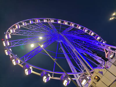 A close-up, low-angle shot captures a Ferris wheel at night in Rimini, Italy. The structure is illuminated with vibrant blue lights along its spokes and supports, while the gondolas are lit with a warm, yellow light. The dark night sky serves as the backdrop, with a few streetlights visible in the upper right corner. The Ferris wheel appears to be in operation or ready for operation, with multiple gondolas visible, some empty and others possibly occupied. The image focuses on the intricate metal framework of the wheel and the geometric patterns created by the lights. The overall impression is one of a festive and illuminated urban attraction against the darkness of the night.
