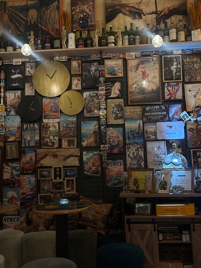 The image shows a wall densely decorated with various items, predominantly framed prints and bottles of alcohol, suggesting a bar or a similar establishment. The prints depict a mix of vintage travel posters for locations like Venice, Rome, Paris, Florence, Malta, and Capri, alongside artwork and some motivational quotes. Some prints feature famous paintings or artistic renditions of people and scenes.
Visible objects include numerous bottles of whiskey and other spirits, as well as decorative items like lamps, small sculptures, and clocks. One notable print displays "The Creation of Adam" by Michelangelo, with the hands almost touching. Another shows a classic image of Marilyn Monroe. There are also signs with text like "Welcome to Haky Cocktail Bar," "I Drink Wine and I Know Things," and "We Do Not Have WiFi."
The overall impression is one of eclectic and dense decoration, with a strong emphasis on travel, classic art, and the theme of drinking. The lighting seems dim, possibly indicating an indoor evening setting. The presence of cocktail bar signage and the variety of alcoholic beverages strongly suggest the location is a bar, potentially in a tourist-friendly area given the travel-themed prints. One of the cropped images includes the text "Valletta, Malta" which confirms the location.