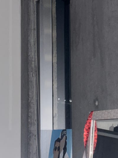 This image depicts an indoor environment in Santa Catarina, Mexico, featuring a vertical composition around a window or glass partition. To the left, a plain light-colored wall or partition is visible. Central to the frame is a dark grey vertical window or door frame with a slender, reflective metallic strip. A light-colored beaded chain, consistent with a blind mechanism, hangs vertically along the left edge of this frame. Through the glass, two distinct bright points of light are reflected against a dark background, suggesting artificial exterior illumination at night or in low light conditions. The right side of the image reveals a dark grey, textured wall with a subtle circular indentation. At the bottom-center, a partial illustration on a light blue background is visible, depicting two stylized human faces, one with sunglasses and another with dark hair and headwear. To the lower-right, a segment of a textured, rectangular object, possibly furniture or a container, is visible, draped with a red fabric patterned with black stars. No individuals are present, nor are there any discernible interactions or actions occurring within the captured scene.