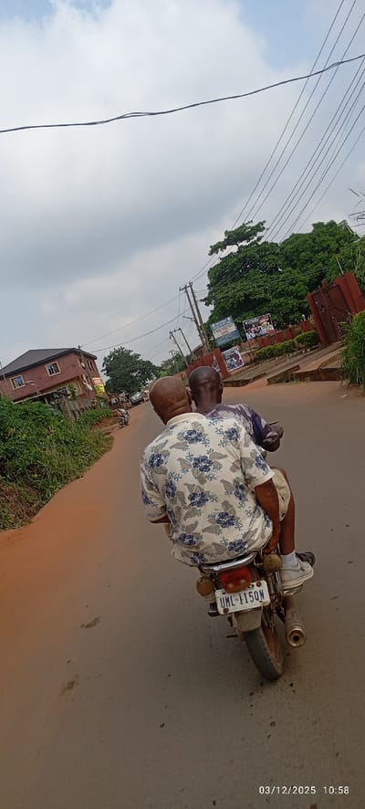 An outdoor scene in Abagana, Nigeria, depicts two adult males riding a motorcycle away from the viewer on a reddish-brown road under a cloudy sky. The passenger, seated behind the driver, is bald and wears a light-colored short-sleeved collared shirt with a blue floral pattern. He is wearing light-colored sneakers with white soles. The driver, seated in front, wears a darker patterned shirt, with a portion of his left arm visible. The motorcycle displays a rear license plate with "UML-115QN" printed in black text on a white background. The bike features a red taillight, amber turn signals, and a visible exhaust pipe on the right side.
To the left of the road, a strip of overgrown vegetation runs alongside, followed by a multi-story reddish-brown building with a dark roof and several windows. Further down the road on the left, another motorcycle is visible in the distance. On the right side of the road, multiple wooden utility poles with numerous overhead wires extend into the background. Behind these poles and a dark brown metal gate, lush green foliage from trees is visible. Two rectangular signs are mounted on a wall or fence on the right; one is blue and white, partially legible as "ST. ANNE CHILD," and the other features graphics and text. The sky is largely overcast with patches of brighter white and light blue. The timestamp "03/12/2025 10:58" is visible in the lower right corner.