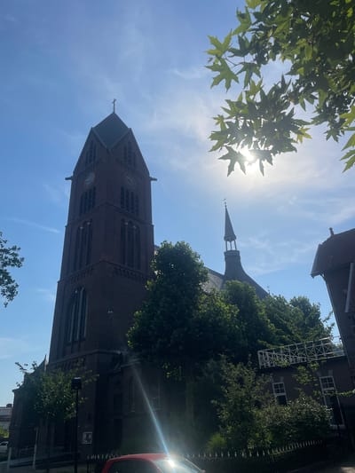 The image depicts a view looking upwards towards the sky, with a brick church tower dominating the frame. The tower has a tall, pointed spire topped with a cross. The church is made of dark brick and features arched windows and clock faces. Lush green trees partially obscure the view of the church, their leaves dappled with sunlight.
The sun is shining brightly, creating a lens flare effect and casting rays across the sky. The sky is a clear blue with some wispy white clouds. The overall impression is one of a bright, sunny day.
The image appears to be taken outdoors in an urban or semi-urban setting, likely in Venlo, Netherlands, given the architectural style of the church. The presence of trees and the clear sky suggest a pleasant daytime scene, possibly late morning or early afternoon. There are no people visible in the image. The image's perspective and the angle of the sun suggest it was taken in the summer months.
