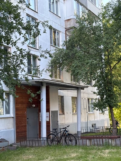 The image depicts the exterior of a multi-story apartment building in Sarov, Russia. The building is constructed with a white, textured facade and features multiple windows, some with decorative grilles. In front of the building, there is a covered entrance area with wooden paneling and a grey door. Two bicycles are parked near the entrance, and a low metal fence encloses a patch of grass. A bench made of concrete and wood is also visible behind the fence. The scene is partially obscured by the branches of leafy trees. The overall impression is that of a residential area in a Russian city.