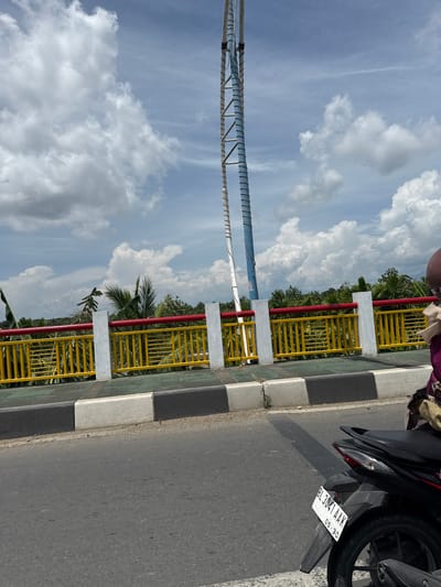 In Lambaro, Indonesia, a street view captures a bright, overcast day. The foreground features the back of a black motorcycle with license plate BL 3041 AAK, suggesting it is parked or stopped near the edge of the road. The motorcycle is partially obscured by the camera's perspective, but its taillight and rear wheel are visible. Beyond the motorcycle, a road surface is evident, marked by a concrete curb with black and white segments.
The middle ground is dominated by a bridge or overpass with a bright yellow metal railing, topped by a red bar. White pillars support the railing at intervals. Behind the railing, lush green foliage, including palm trees, creates a natural backdrop. A tall, thin structure, possibly a telecommunications tower or utility pole, with a blue and white segmented shaft, stands prominently against the sky.
The sky is filled with large, fluffy white clouds against a light blue canvas, indicating a dynamic weather pattern. The overall scene depicts a typical day in an Indonesian town, with infrastructure and natural elements coexisting.
