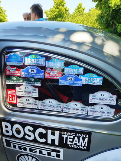 The image shows the rear passenger side window of a silver car, adorned with numerous stickers for various car rallies and events. The stickers include "Silvretta Classic 2018," "Ramberg Historic" (multiple years), "ADAC-Bayerwald-Rallye" (multiple years), "DMI Histo Day+Night Challenge," and "Donau Classic" (multiple years). A "Gloria Feuerlöscher" (fire extinguisher) sticker is also visible. The car is parked outdoors, with bright sunlight casting reflections on its surface. Green trees and a bright blue sky are visible in the background. Two men are partially visible through the window, looking into the car. One man has gray hair and is wearing glasses, while the other has dark hair and is wearing sunglasses. Below the window, a large "BOSCH" sticker and a "PIRELLI" sticker are prominently displayed on the car's bodywork, along with a "RACING TEAM VIENNA" sticker. The image appears to have been taken in Pirmasens, Germany, as indicated by a sticker reading "Pirmasens" on one of the rally decals.