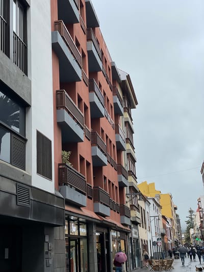 The image shows a street view in San Cristóbal De La Laguna, Spain, focusing on a modern building facade. The building is characterized by a reddish-brown exterior with projecting, angular gray balconies and wooden lattice screens. The lower portion of the building is white with rectangular ventilation grates. The building rises up and along the right edge a street is seen with people walking under umbrellas. Various shops with large glass windows are visible, and the facades of other buildings line the street. The sky is overcast.