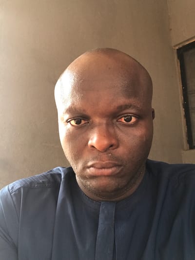 An adult male is depicted in a frontal head-and-shoulders portrait, positioned against a plain, light brown wall in an indoor setting in Egboje, Nigeria. The subject, of African descent, is bald with dark brown skin. He wears a dark blue garment, consistent with a tunic, featuring a mandarin collar and a central placket; subtle vertical stitching is visible on the upper left shoulder area. His dark eyes present with a reddish tint in the sclera, and minimal stubble is visible across his chin and jawline. He maintains a direct, neutral gaze towards the observer. A dark-framed aperture, possibly a window or doorway, is partially visible in the upper right background.