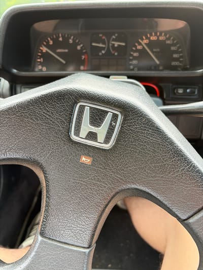 The image shows a close-up of a car's dashboard and steering wheel, with a Honda logo prominently displayed on the steering wheel. The speedometer indicates a speed of approximately 80 km/h. The tachometer is also visible, showing an engine speed of around 2000 RPM. A small orange icon resembling a horn is visible on the steering wheel. The scene appears to be indoors, possibly in a garage, as indicated by the lighting and the absence of any outdoor elements. There is no indication of the car's location in Kumachevo, Russia.