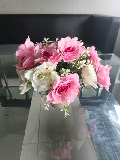 The image displays a close-up, eye-level view of an artificial flower arrangement situated on a reflective glass-top table within an indoor setting. The bouquet primarily consists of light pink and white faux roses, interspersed with small white filler flowers and green artificial foliage. The flowers are arranged in a cylindrical pot, which appears to be covered with a textured, light brown material, possibly burlap or a similar fabric.
The glass surface of the table reflects the base of the pot, the lower parts of the flowers, and parts of the room's environment, including the tiled floor below the table. The flooring visible beneath the table features light-colored, rectangular tiles. In the blurred background, a dark, indistinct object, likely a piece of furniture such as a sofa or cabinet, occupies the upper portion of the frame. The lighting suggests an interior space. No individuals, actions, or interactions are depicted. While the prompt specifies the location as the Special Capital Region of Jakarta, Indonesia, no discernible geographical markers within the image itself confirm this specific location.