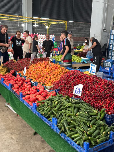 The image depicts an active indoor fruit and vegetable market located in Edirne Merkez, Turkey. The scene features a large, well-stocked stall displaying various fresh produce. The floor is concrete, and the background consists of a dark grey brick-patterned wall with black metal roller shutters at the top, along with a grey concrete pillar on the right. Yellow metal support beams run across the top, from which several bare light bulbs hang, providing illumination.

In the foreground, a prominent display table is covered with green artificial turf, holding numerous blue plastic crates filled with produce. From left to right, these include large piles of red strawberries, yellow bananas, large quantities of bright orange apricots, several crates of red tomatoes, and substantial mounds of dark red cherries. A pile of green cucumbers is visible in the lower right corner. White price signs are interspersed among the produce; one sign for cherries reads "60" and another, further back, indicates "Kilo 120."

Multiple individuals are present, engaging in market activities. On the left, a man in a dark t-shirt and camouflage shoulder bag holds a clear plastic bag containing orange items, possibly carrots. Next to him, a woman in a black t-shirt with a white butterfly graphic observes the stall. Behind her, a man in a beige polo shirt appears to be conversing. To the right of the woman, a young male vendor wearing a black t-shirt with "MONCLER" text on the sleeve and a green apron stands behind the stall, looking toward the left. Further to the right, an older man in a blue baseball cap is visible behind the stall. In the middle-right, another man in a black t-shirt is seen placing items into a light blue plastic bag, suggesting a purchase. Several other customers and vendors are visible in the background, including a small child near a display of green plums.

The grey pillar on the right side of the image has handwritten text on it, including "HASIR," "ÖZKAN B," and "MEHME," likely names or parts of business names in Turkish. In the far background, behind the main produce stalls, stacks of red and white Persil brand detergent bottles are visible, indicating that other goods are also sold in this market area. A blue weighing scale is present on a table further back, behind the cherry displays.