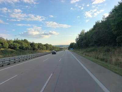The image depicts a view from a vehicle traveling on a highway, likely an Autobahn, in or near Frankenberg/Sachsen, Germany. The highway has multiple lanes separated by solid white lines and a concrete barrier on the left side. The road surface is asphalt. Green trees and vegetation line both sides of the highway, with a dense forest visible on the right. The sky is blue with scattered white clouds, suggesting a pleasant, possibly late afternoon, weather condition. Several cars are visible on the highway, including a dark-colored SUV in the left lane and two other vehicles further ahead. The presence of road signs and the general infrastructure indicates a European highway. The terrain appears to be gently rolling hills in the distance.