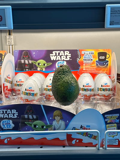 This image depicts a retail display in a store in Jestetten, Germany. The primary subject is a dark green, textured avocado centrally positioned within a clear plastic tray designed to hold confectionary eggs. Surrounding the avocado are multiple white, foil-wrapped Kinder Überraschung (Kinder Surprise) eggs, arranged upright in the tray's compartments.
Behind and beneath the eggs, Star Wars-themed promotional cardboard material is visible. The top banner features the "STAR WARS™" logo, text "JETZT IN JEDEM", and cartoon illustrations of Darth Vader and Grogu. It also advertises a "GEWINNCHANCE 1 von 1.111 STAR WARS TASSEN" (chance to win 1 of 1,111 Star Wars mugs), showing an image of a black Star Wars mug. The bottom banner similarly displays the "STAR WARS™" logo, text "JETZT IN JEDEM 7. EI!", and cartoon illustrations of Darth Vader, Grogu, and Luke Skywalker holding a blue lightsaber.
The environment includes a dark blue shelf or structure above the display. A white price tag on the left shows "9" and "q - 59.50". A smaller blue price tag on the right details "Alesto Snack2go Nu...", "geröstet & ge...", and "50 g Beutel", alongside a QR code and barcode. A white vertical element with "GS", "VDE", and "CE" markings is positioned between the price tags. The display rests on a lower shelf, partially obscured by a white metal guard rail in the foreground. No people are visible.