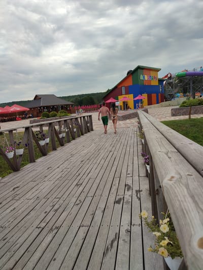 A couple walks on a wooden boardwalk at an outdoor water park in Shcherbani, Ukraine. The man is wearing green swim shorts and the woman is wearing a black bikini. They are walking away from the camera, towards a brightly colored building with a sign that reads "АКВАПАРК". To the left of the boardwalk are red umbrellas and tables, suggesting a dining or seating area. In the background, there are water slides. The sky is overcast with gray clouds. The boardwalk is lined with wooden railings and potted flowers. The wooden planks of the boardwalk show water stains and footprints.