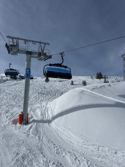 The image depicts a snowy mountain landscape at a ski resort in Saint-Martin-de-Belleville, France, under a partly cloudy sky. A gray metallic ski lift pylon, labeled "4/13" and featuring an attached blue informational sign, stands prominently in the mid-ground, secured at its base by orange protective padding. A steel cable extends horizontally from the pylon, supporting multiple empty blue and black ski lift chairs. One chair is visible to the immediate right of the pylon, and others are further along the cable to the left and in the distance. The snow-covered ground displays numerous ski tracks, particularly on the slope behind the pylon. Scattered evergreen trees are visible on the distant snow-covered hills. A distinct snow feature, possibly a terrain park element, is in the lower right foreground. The pylon casts a long, distinct shadow across the snow. No individuals are present in the scene.