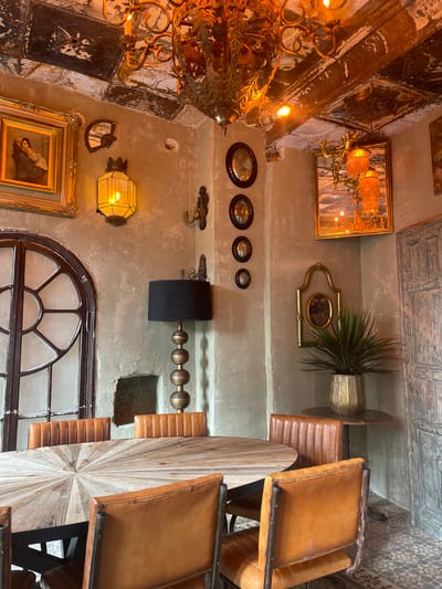 The image depicts an interior view in Jerez De La Frontera, Spain. The setting appears to be within a restaurant or bar, indicated by the presence of tables and leather-upholstered chairs. The decor is eclectic, including an antique chandelier, framed paintings, and ornamental wall decorations. Architectural elements include a segmented arched window and a textured ceiling. The lighting is a mix of natural and artificial, with lamps and a window providing illumination.