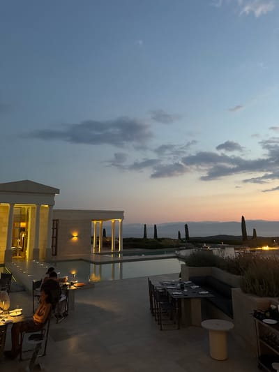 The image shows an outdoor dining area at dusk in the Municipal Unit of Kranidi, Greece. A sleek infinity pool with a modern architectural structure featuring classical columns is visible to the left, illuminated by warm interior lighting. Several tables are set for dining, with plates, glasses, and cutlery arranged on them. Diners are seated at some of the tables, with one woman in a sparkly dress in the foreground appearing to be dining alone. In the background, a serene landscape with distant mountains and a body of water can be seen under a twilight sky with scattered clouds. Tall, slender trees punctuate the horizon. The overall atmosphere suggests a luxurious and tranquil dining experience.