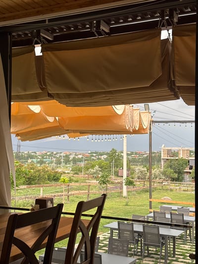 The image shows an outdoor seating area with tables and chairs, likely a cafe or restaurant, set against a backdrop of a rural landscape. The foreground is dominated by rows of folded beige fabric awnings or curtains, suggesting a shaded area. Behind these, glimpses of outdoor dining furniture, including dark wooden chairs and grey metal tables, are visible. Beyond the seating area, a green, grassy field stretches out, interspersed with trees and some low-lying buildings that indicate a settlement or village. In the distance, a body of water is visible under a cloudy, overcast sky, suggesting a temperate or possibly cool day. The overall impression is one of a tranquil, open-air venue. There is no visible text in the image.