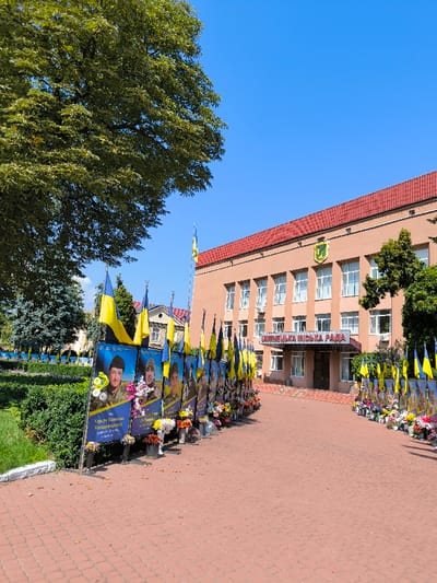 An outdoor, eye-level shot captures the Illintsi City Council (ІЛЛІНЕЦЬКА МІСЬКА РАДА) building in Illintsi, Ukraine, under a clear blue sky. The building features a light peach-colored facade, multiple windows, a red tiled roof, and the city's coat of arms above the entrance. A wide, reddish-orange paved pathway leads directly to the building's entrance.
Flanking both sides of this pathway is a double-row arrangement of vertical memorial banners. Each banner displays a portrait photograph, primarily of men in military attire, with Ukrainian text below the image. A Ukrainian national flag (blue and yellow) is attached to the top of each banner. At the base of each banner, various floral arrangements, consisting of both fresh and artificial flowers in buckets and vases, are positioned on the ground.
To the left of the pathway, a large, mature tree with dense green foliage is prominent, along with manicured green bushes and several evergreen trees. The scene is brightly illuminated by direct sunlight.