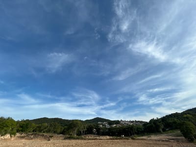 The image depicts a landscape in Baan Wang Ta Kien, Thailand, under a partly cloudy blue sky. The ground is a barren, earthy brown, with evidence of excavation. A line of trees separates the foreground from a hillside densely covered with lush green vegetation. Residences are visible on the hillside, nestled among the trees. The sky is mostly blue with wispy, cirrus-like clouds spread throughout.