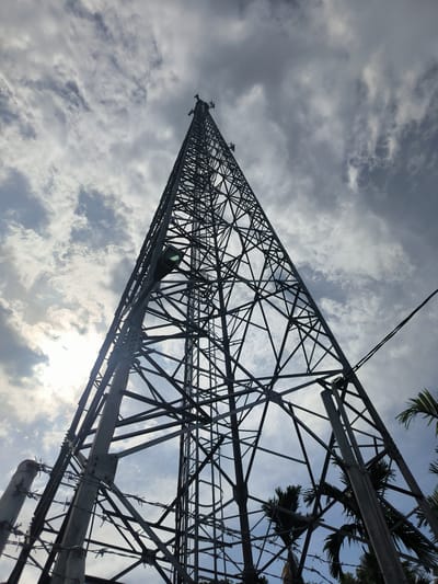 This media file features a low-angle shot looking up at a tall, lattice-style telecommunications tower against a cloudy sky. The tower is the primary subject, constructed from dark metal beams forming a strong, geometric structure that tapers towards the top. Several antennas are visible at the apex of the tower. A light fixture is attached to one of the tower's vertical supports, positioned midway up the visible portion. The lower foreground shows a section of barbed wire fence, suggesting the tower is in an enclosed or secured area. Palm fronds are visible on the right side, hinting at a tropical or subtropical location. The sky is filled with dramatic, grey and white clouds, with patches of bright sunlight breaking through, indicating a dynamic weather condition and likely late afternoon or early morning. The overall mood is somewhat stark and industrial, juxtaposed with the natural elements of the sky and palm trees. No people are visible, and there is no visible text in the image. The setting is identifiable as Cotmerbo, Indonesia.