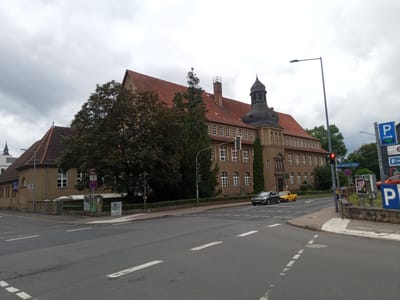 The image captures a street view in Eisenach, Germany, near a large building that appears to be a school or institutional facility. The building has a distinctive red-tiled roof, a prominent tower with a black, conical roof, and many regularly spaced windows. A tree with dense foliage stands in front of the building. Several cars are visible on the street, including a dark sedan and a yellow pickup truck. Traffic signals and street signs, including a parking sign with "FREI" (free) indicated, are present. The sky is overcast, suggesting cloudy weather. The foreground shows a road with lane markings and a pedestrian crossing.