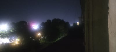 The image captures a nighttime urban scene in Andijan City, Uzbekistan, viewed from a high vantage point, possibly a balcony or window. The foreground is dominated by the textured surface of a wall on the right side. Beyond this, dark foliage, likely trees, obscures much of the mid-ground. In the distance, city lights are visible, illuminating the night sky with bright, concentrated sources of light. Several buildings are discernible, with some appearing to be residential or office blocks. The overall atmosphere is one of a dimly lit, expansive urban landscape at night.