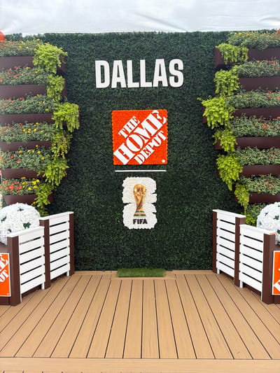This media file shows an outdoor display, possibly at an event or promotional setup, in Dallas, USA. The primary subject is a large, artificial green hedge wall that serves as a backdrop. Prominently featured on the wall are large, white, block letters spelling "DALLAS" vertically. To the right of the text, there are two decorative elements. The first is an orange, square-shaped logo with white text that reads "THE HOME DEPOT". Adjacent to this is a white floral arrangement shaped like a trophy, with "FIFA" written on it in blue letters.
In front of the hedge wall, there are planters filled with lush green foliage and some small yellow and orange flowers, adding a touch of nature to the scene. To the right of the hedge wall, a wooden deck extends out. On the deck, there are two white benches with brown trim, positioned facing the hedge wall. White floral arrangements resembling large pom-poms are placed on either side of the benches.
The lighting suggests it's daytime, and the setting appears to be a well-lit outdoor area. The overall impression is that of a vibrant and engaging display, likely for a public event or a sponsored activation. There is no visible indication of people actively participating in an event, making it seem like a staged setup. The wooden decking, artificial greenery, and the clear logos suggest a commercial or event-related context.