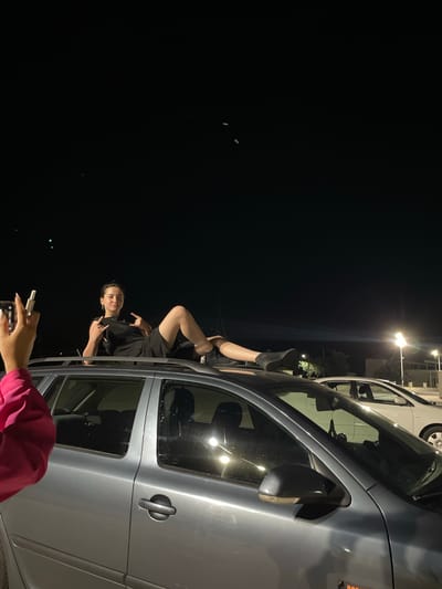 In Limenas Avdiron, Greece, at night, a person lies on the roof of a silver station wagon, their legs extended and feet resting on the edge. The individual is wearing a black t-shirt and black shorts, with black sneakers. Their right hand is raised, fingers forming a rock-on gesture, while their left hand is also raised, similarly posed. To the left of the frame, a person's left arm, clad in a bright pink garment, is visible, holding a smartphone up, seemingly to take a photo. The background is dark, with the night sky above and some faint lights and structures in the distance, including streetlights and the headlights of another car. The station wagon appears to be parked outdoors, possibly in a lot or on a street.