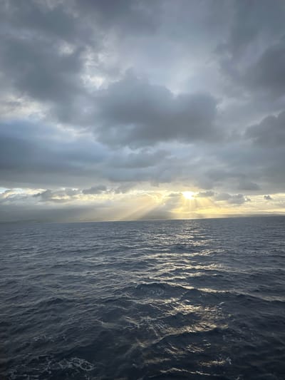 The image shows a view of the ocean from a boat, with a cloudy sky and sunlight breaking through the clouds. The ocean is choppy, with waves reflecting the sunlight. In the distance, a landmass is visible, likely the coast of Guadeloupe. The scene appears to be during either sunrise or sunset, given the angle of the sunbeams. There are no people or specific objects visible in the foreground or on the water. The overall impression is one of natural beauty and the power of the elements.