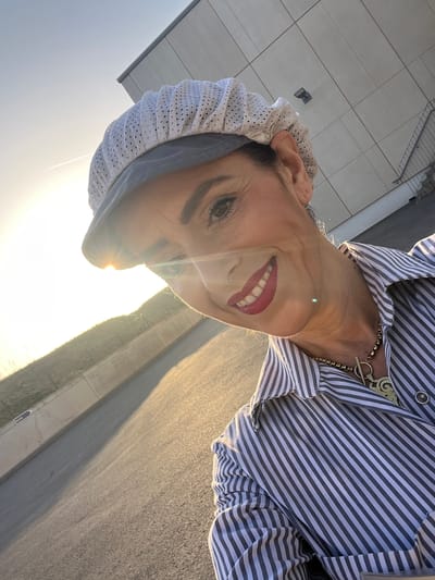 The image shows a woman in Mensdorf, Luxembourg, wearing a white perforated hair covering and a vertically striped shirt with a gold-colored necklace. The background includes a street with sidewalks and signage. The sunlight appears strong.