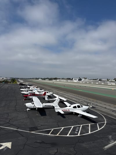 This aerial image depicts a row of small airplanes parked on an asphalt surface, likely a runway or tarmac, at an airport. The aircraft are predominantly white, with some featuring red accents. Visible text on the planes includes registration numbers like "N258JS", "N5000Y", and "N724TT", along with model designations such as "SR20". The ground is marked with white lines, including a circular "NO PARKING" stencil, and numerical markings like "3165".

In the background, a long, straight runway or taxiway stretches into the distance, bordered by green and grey strips. Buildings with white facades and trees are visible further beyond the runway. The sky is a mix of blue and overcast clouds, suggesting a partly cloudy day. The lighting indicates it is daytime, possibly late morning or early afternoon, given the shadows cast by the aircraft. The scene appears to be set in a location with clear weather and good visibility, typical for an airport environment. The overall impression is one of organized aviation infrastructure and readiness.