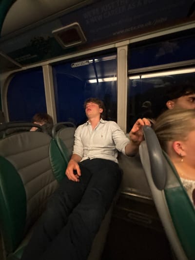 The image shows a person sleeping across two seats on public transportation, possibly a bus or train. The individual is wearing a light-colored, button-down shirt and dark pants. Above them, a partially visible individual with blonde hair is seen. The interior of the vehicle is visible with green and gray seating. An advertisement for "A New Career" is visible on the window, along with an emergency exit sign. The picture is taken at an angle, making the scene appear tilted.