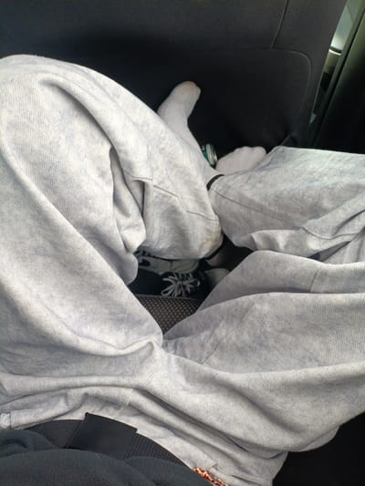The image shows a person sitting in a car. They are wearing grey sweatpants and white socks. Their shoes are visible beneath the sweatpants. A can is visible on the seat next to them. A seatbelt is buckled across their lap. The car has black cloth seats. The image provides no evidence to indicate this photo was taken in Malaya Kuskunka, Russia.