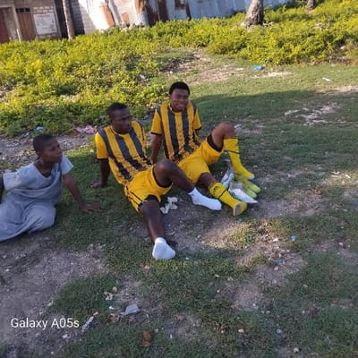 Three men are sitting on the ground, two in yellow and black striped soccer uniforms and one in a gray shirt. The two men in uniforms are wearing white socks. The men are sitting on a patch of grass in Nungwi, Tanzania, at the coordinates -5.726 latitude, 39.301 longitude. They appear to be in a relaxed mood. To the left of the men, a young boy in a gray shirt is sitting on the grass.