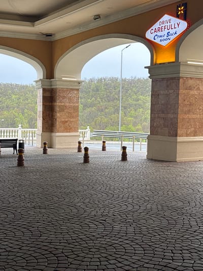 The image depicts a covered walkway with decorative columns and arches, leading towards a roadway with a metal barrier and a distant view of greenery, possibly trees. The walkway is paved with a dark gray cobblestone pattern in an arched design. The columns are clad in marble with beige or cream-colored accents, and the arches are finished in a similar light tone.
Along the walkway are a series of dark brown bollards with yellow stripes. A black bench is visible in the lower portion of the image, between two of the bollards.
To the left of the frame is a vintage-style sign with the words "DRIVE CAREFULLY Come Back SOON" in a classic font, indicating a place or destination. A starburst emblem is above the text.
The overall scene suggests a location with architectural features and a pathway leading out of an enclosed area towards a road or highway. The setting could be Shkotovo, Russia.