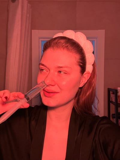 An adult female is observed in an interior room located in Nikol'skii, Russia. She is performing a nasal irrigation procedure, holding a clear plastic nasal irrigator with a plunger to her right nostril. The device contains a clear liquid with blue volume markings. The woman has light skin, brown hair styled away from her face, and wears a light-colored, textured headband. She is dressed in a dark, possibly black, satin or silk robe. Her left hand, featuring red nail polish on the thumb, grips the irrigator. Her gaze is directed towards the upper right, and her mouth is slightly open. The scene is illuminated by a prominent reddish-orange light. The background includes light-colored walls, a white framed doorway or opening with a blue hue visible through it, a white curtain on the left side, and a dark shelving unit holding baskets on the right.