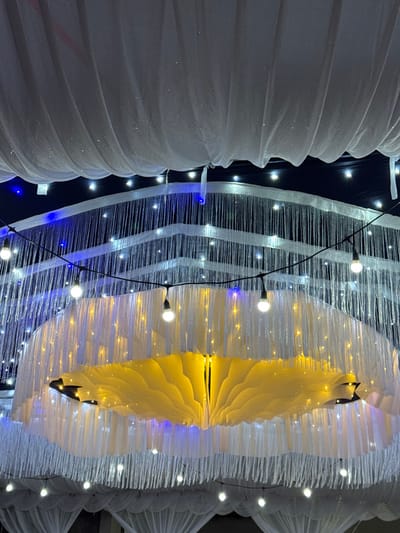 The scene captures an illuminated decorative canopy within an event space, identified as being in Cuôr Đăng B, Vietnam. No individuals are present. The overhead structure features layers of suspended elements. The uppermost layer consists of draped white, translucent fabric with fine glitter particles. Below this, multiple tiered sections are formed by hanging white, shimmering thread-like strands, illuminated by discreet cool white and blue lights from behind. A black string light fixture, strung horizontally, displays exposed filament bulbs emitting warm white light. Dominating the central area is a large, inverted dome-shaped fixture constructed from layered white fabric. Its interior glows with a bright yellow-orange light, creating a ruffled, petal-like texture. Additional fine white strands hang from the periphery of this central fixture. The lower background shows further installations of gathered white fabric drapes. The environment is dark, suggesting an indoor setting or nighttime event, with all illumination sourced from artificial lighting elements.