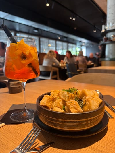 The scene depicts a dining table within a restaurant located in Kiryat Ono, Israel. In the foreground, a stemmed glass holds an orange-hued beverage with ice, numerous orange slices, and two black straws, resting on a black square coaster on a light-colored wooden table. Adjacent to it, a dark brown, ribbed ceramic bowl, seated on a circular dark plate, contains multiple golden-brown, fried rectangular food items, garnished with finely chopped green herbs and thin strips of dark seaweed. A silver spoon is present in the bowl, and a pair of wooden chopsticks is partially visible to the right of the bowl. A silver fork and knife are positioned on the table surface in the bottom left corner. The background, largely blurred, shows the interior of a contemporary restaurant with a dark ceiling, recessed lighting, and large windows featuring horizontal blinds or grates. Several indistinct figures of individuals are seated at tables throughout the restaurant, engaged in dining or social interaction. A decorative, multi-tiered structural element is partially visible on the right side of the frame.