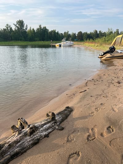 The image depicts a serene lakeside scene, likely at dusk given the soft lighting. The foreground is dominated by a sandy beach with visible footprints, suggesting recent human activity. A weathered piece of driftwood lies prominently on the sand.

In the water, a small white boat is moored near the shore, and further back, a larger inflatable boat with an outboard motor is pulled up onto the beach, partially covered by a yellow tarp. The shoreline is lined with lush green trees and foliage, creating a natural backdrop. The water itself is calm, reflecting the overcast sky.

A single person is visible near the white boat, standing on a structure extending into the water, but their activity is unclear. The overall impression is one of a peaceful, natural setting, possibly a recreational area. The presence of boats and a beach suggests a location for outdoor leisure activities. There is no discernible text in the image.