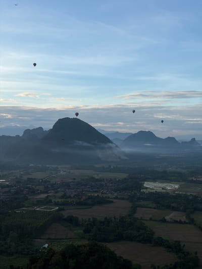 This is a high-angle, full shot of a landscape in Naka, Laos, dominated by rolling hills and mist. In the sky, several hot air balloons are visible, adding a splash of color and activity to the scene. One balloon, colored red, is positioned near the peak of a prominent green hill. Another balloon with stripes of various colors floats in the upper left quadrant of the image, while a blue and white striped balloon is seen in the middle right. A dark-colored balloon is also visible higher up in the sky. The landscape below is a patchwork of green forests, rice paddies, and scattered buildings, suggesting a rural setting. The presence of mist at the base of the hills and in the valleys contributes to a serene and slightly mysterious atmosphere. A dragonfly is captured in flight near the top left of the frame.