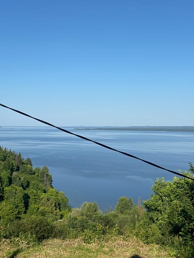 A wide-angle, eye-level shot captures a serene natural landscape in Kostovaty, Russia, under a clear, bright blue sky. The foreground is a grassy slope dotted with dry grass and low-lying green bushes, with a dark, diagonal power line cutting across the upper portion of the frame. Beyond the slope, a vast, tranquil blue body of water stretches towards a distant, hazy shoreline lined with a dense forest of green trees. The water exhibits subtle ripples, suggesting a gentle breeze. A small, dark green object, possibly a boat, is visible on the water in the mid-ground. The overall scene conveys a sense of peaceful isolation and natural beauty.