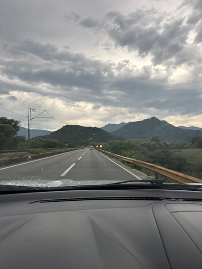 The image depicts a view from inside a vehicle traveling on a two-lane road, likely in or near Braćeni, Montenegro. The road is flanked by a rusty guardrail on the right and a gentle slope with trees and vegetation on the left. In the distance, a series of verdant mountains rise under a dramatic, cloudy sky. Power lines are visible overhead, supported by utility poles. A few vehicles can be seen on the road ahead, including a white car and a vehicle with flashing orange lights, suggesting a possible hazard or work zone. The dashboard of the vehicle is partially visible in the foreground. The overall atmosphere is overcast, with the clouds suggesting the possibility of rain or a recent shower.