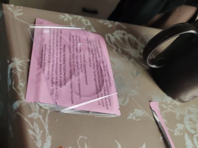 A pink sheet mask packet lies on a patterned tablecloth. The packet is partially opened, revealing a clear plastic backing. Text in Russian and English is visible on the packet. The Russian text, located on the left side, provides instructions and warnings. The English text, on the right, lists ingredients such as "Water, glycerin, propylene glycol, beta-glucan, ceratonia siliqua gum, hydrogenated castor oil, chlorophenyl ether, hydrogenated dmdm hydantoin, essence." A dark mug with a curved handle is visible to the right of the packet. The lighting suggests an indoor environment, possibly a domestic setting. The overall scene is static and shows no signs of activity or interaction. No specific location in Pioner, Russia can be determined from the image.