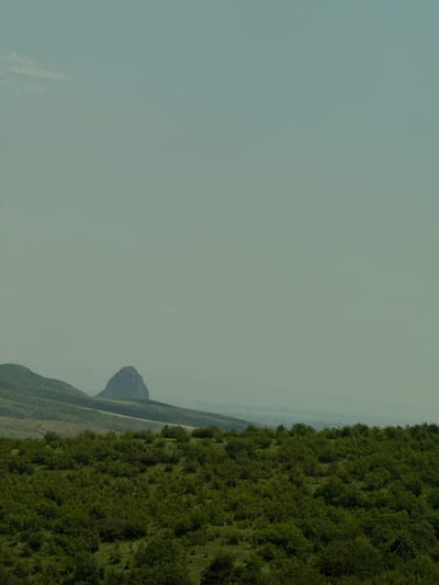 This is an outdoor, eye-level shot of a slightly hazy landscape. In the foreground, a dense, green forest of trees and bushes stretches across the frame. Beyond the forest, rolling hills with sparse vegetation are visible, leading to a more distant, conical rock formation. The sky is a pale, muted blue with a faint haze. The scene appears to be a natural, undeveloped area, possibly in the region of Voskepar, Armenia. There are no people or animals present.